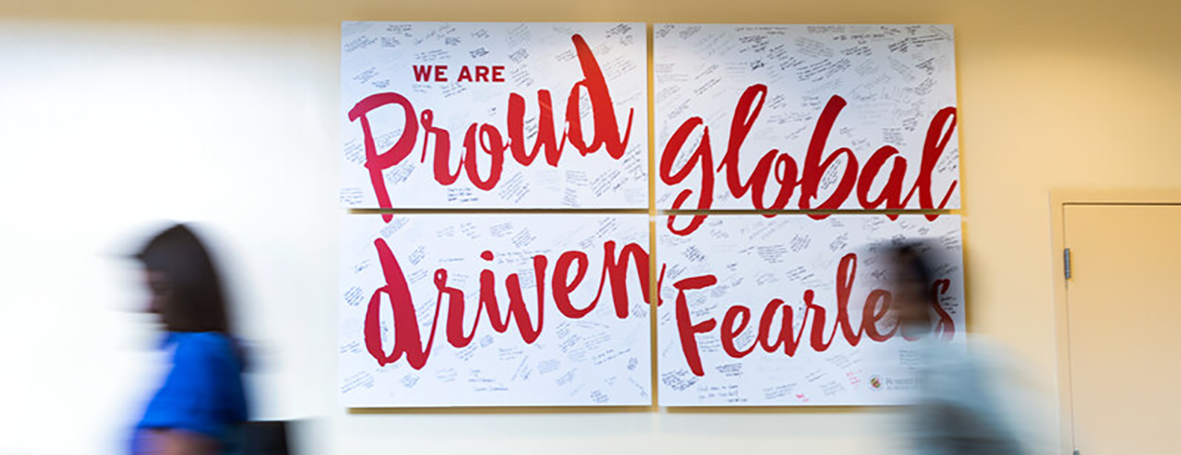 Students passing by a poster on the wall that reads 'We are proud, global, driven, fearless'